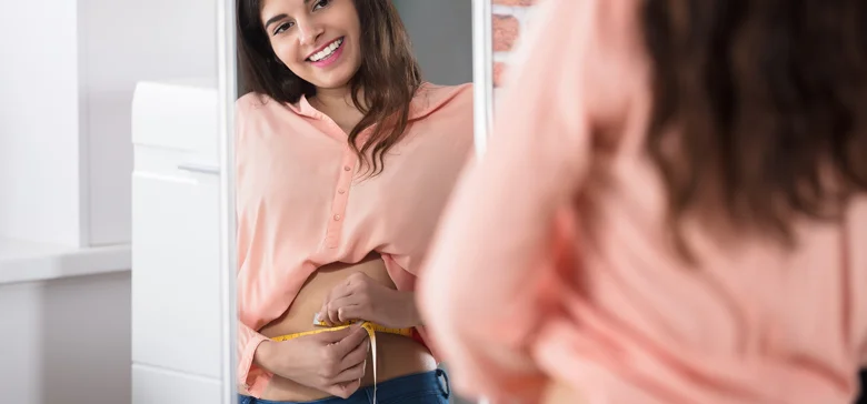 Frau schaut Bauch in Spiegel an mit Messband