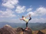 Katharina beim Yoga auf einem Felsen in den Bergen, führt eine Balance-Pose aus. Im Hintergrund klare blaue Himmel und hügelige Landschaft.